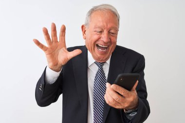 Üst düzey gri saçlı işadamı, izole edilmiş beyaz arka planda duran akıllı telefonu kullanıyor. Çok mutlu ve heyecanlı. Kazanan ifade zaferi büyük bir gülümsemeyle kutluyor ve ellerini kaldırıyor.