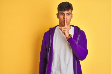 Mor kazak giyen genç Hintli adam izole edilmiş sarı arka planda dikiliyor ve dudakları parmaklarıyla sessiz olmak istiyor. Sessizlik ve gizli konsept.