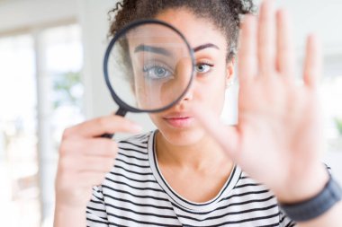 Genç Afro-Amerikalı kadın büyüteçle bakıyor, eli açık, dur işareti yapıyor, ciddi ve kendinden emin bir ifadeyle, savunma hareketi yapıyor.