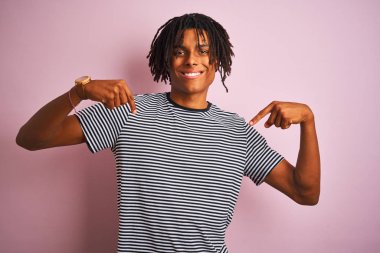 Sarı çizgili tişört giyen Afro bir adam izole edilmiş pembe arka planda duruyor. Yüzünde bir gülümsemeyle, gururlu ve mutlu bir şekilde kendini işaret ediyor..