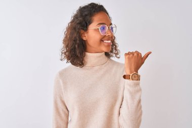 Balıkçı yaka kazak ve gözlük takan genç Brezilyalı bir kadın izole edilmiş beyaz arka plan üzerine işaret ediyor ve gülümseyen bir yüzle baş parmağıyla onu gösteriyor.