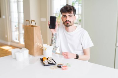 Eve servis yaparken Asya suşisi yiyen ve akıllı telefon ekranı gösteren akıllı yüzlü, ciddi düşünen genç bir adam.