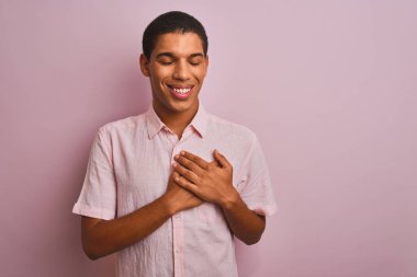 Soyutlanmış pembe arka plan üzerinde duran, elleri göğsünde, gözleri kapalı ve yüzünde minnet dolu bir jest olan genç, yakışıklı Arap bir adam. Sağlık kavramı.