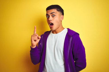Mor kazak giyen genç Brezilyalı adam izole edilmiş sarı arka planda duruyor ve başarılı bir fikirle işaret ediyor. Heyecanlı ve mutlu. Bir numara..
