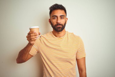 Genç Hintli bir adam, izole edilmiş beyaz arka planda duran bir fincan kahve içiyor. Zeki bir yüz ifadesiyle ciddi ciddi düşünüyor.