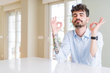 Genç iş adamı beyaz masada oturuyor rahatlıyor ve gözleri kapalı gülümsüyor parmaklarıyla meditasyon hareketi yapıyor. Yoga konsepti.