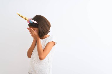 Tek boynuzlu at diademi takan güzel bir kız çocuğu, izole edilmiş beyaz arka planda dikiliyor, üzgün bir ifadeyle elleri ile yüzünü kapatıyor ve ağlıyor. Depresyon kavramı.