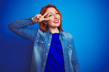 Mavi izole arka plan üzerinde duran denim gömlek giyen genç güzel kızıl saçlı kadın yüzü üzerinde parmakları ile barış sembolü yapıyor, zafer gösteren neşeli gülümseyen