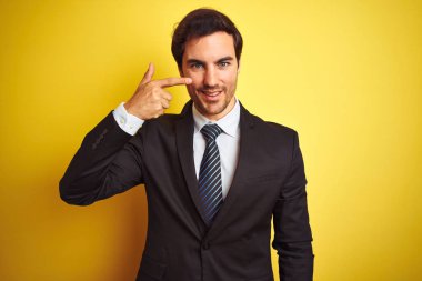 Takım elbiseli ve kravatlı genç yakışıklı bir işadamı izole edilmiş sarı arka planda duruyor. El parmağı ve burunla işaret ediyor, neşeli bir şekilde gülümsüyor. Güzellik konsepti