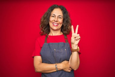 Kırmızı izole edilmiş arka planda önlük giyen orta yaşlı bir kadın kameraya gülümseyip parmaklarıyla zafer işareti yapıyor. İki numara..