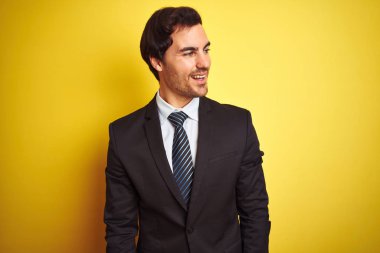 Takım elbiseli ve kravatlı genç yakışıklı bir işadamı izole edilmiş sarı arka planda durup yüzünde gülümseme, doğal bir ifade ile yan yana bakıyor. Kendinden emin gülen.