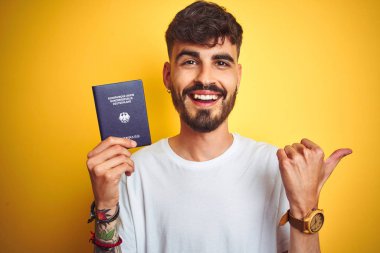 Alman pasaportu takan genç bir adam izole edilmiş sarı arka planda ve baş parmağıyla gülümsüyor.