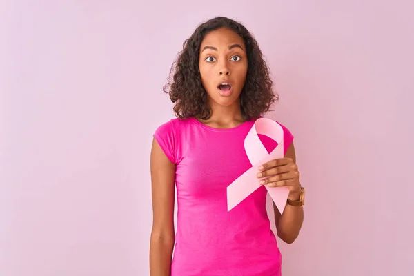 若いブラジル人女性保持癌リボン上に立って隔離されたピンクの背景怖がってショックで驚きの顔 恐れと恐怖の表現で興奮 — ストック写真