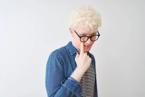 Young albino blond man wearing denim shirt and glasses over isolated white background Pointing to the eye watching you gesture, suspicious expression