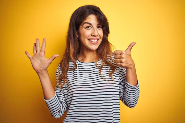 Young beautiful woman wearing stripes t-shirt standing over yelllow isolated background showing and pointing up with fingers number six while smiling confident and happy.