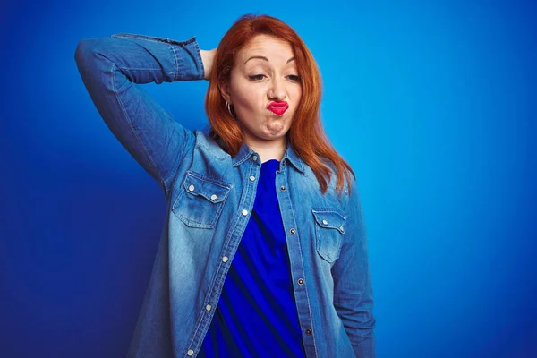 Young beautiful redhead woman wearing denim shirt standing over blue isolated background confuse and wondering about question. Uncertain with doubt, thinking with hand on head. Pensive concept.