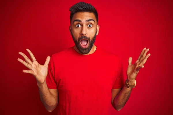 孤立した赤い背景の上にTシャツを着た若いハンサムなインド人男性が狂気を祝い 腕を上げて興奮して叫んで目を開けて成功したことに驚いています 勝者のコンセプト — ストック写真