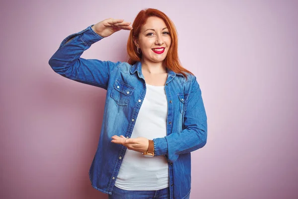 Youg beautiful redhead woman wearing denim shirt standing over isolated pink background gesturing with hands showing big and large size sign, measure symbol. Smiling looking at the camera. Measuring concept.