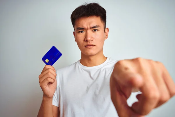 Young asian chinese man holding credit card standing over isolated white background pointing with finger to the camera and to you, hand sign, positive and confident gesture from the front