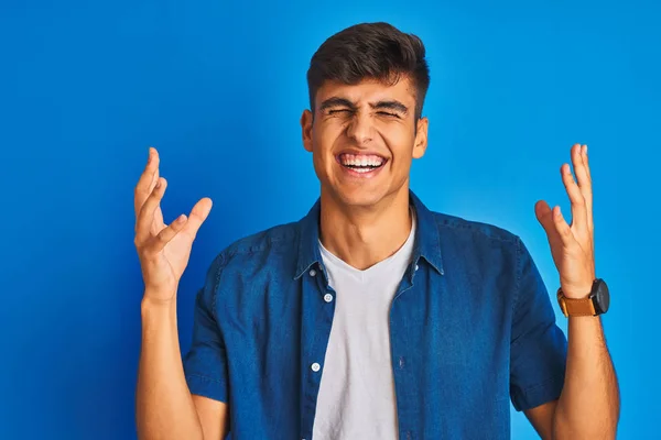 孤立した青い背景の上にシャツを着ている若いインド人男性は 腕を上げて興奮して叫んで目を閉じて成功のために狂気と狂気を祝います 勝者のコンセプト — ストック写真