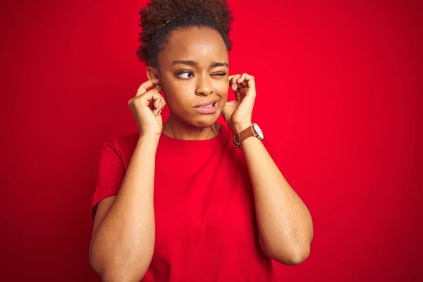 アフリカ系アメリカ人の若い美しい女性は 孤立した赤い背景の上に耳を指で覆い 大きな音楽のノイズのためにいらいらする表現をしています ろう者の概念 — ストック写真