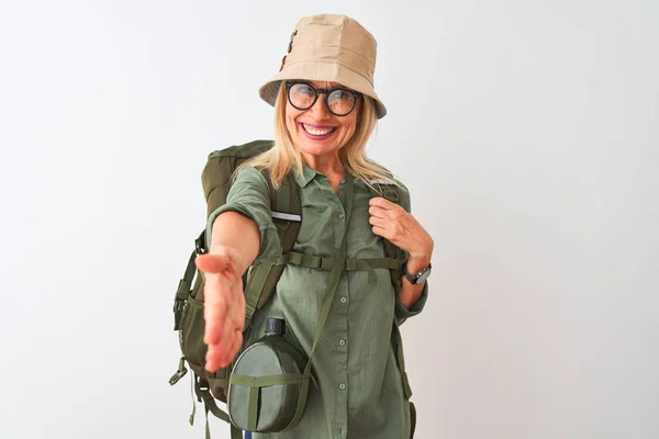 Middle age hiker woman wearing backpack hat canteen glasses over isolated white background smiling friendly offering handshake as greeting and welcoming. Successful business.