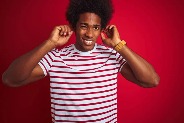 Young african american man with afro hair wearing striped t-shirt over isolated red background covering ears with fingers with annoyed expression for the noise of loud music. Deaf concept.
