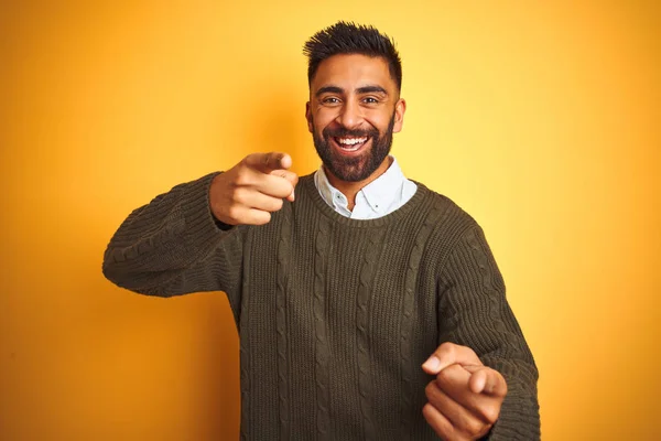 Young indian man wearing green sweater and shirt standing over isolated yellow background pointing fingers to camera with happy and funny face. Good energy and vibes.