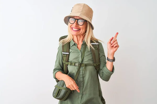 Middle age hiker woman wearing backpack hat canteen glasses over isolated white background with a big smile on face, pointing with hand and finger to the side looking at the camera.