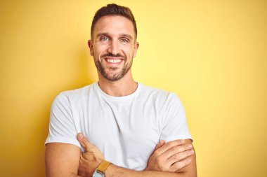 Sarı izole edilmiş arka plan üzerine sıradan beyaz tişört giyen genç yakışıklı adam kameraya bakarken kollarını kavuşturup gülümsüyor. Pozitif kişi.
