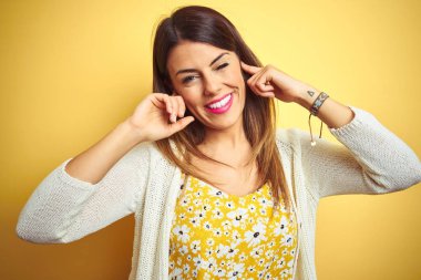 Sarı arka plan üzerinde duran ceketli genç bir kadın yüksek sesli müzik sesi için kulakları parmaklarıyla kapatıyor. Sağır kavramı.