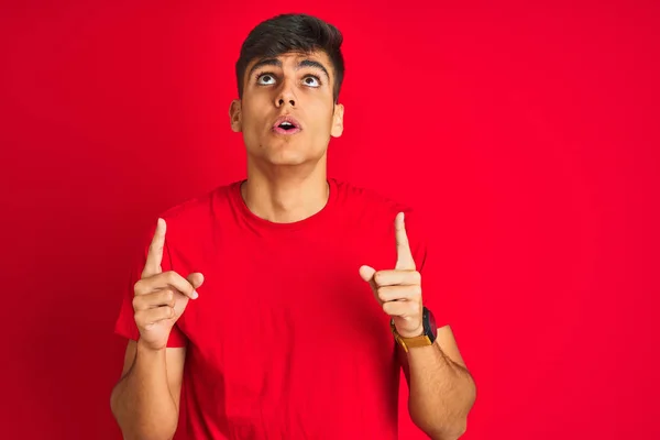 穿着T恤衫站在孤立的红色背景上的印度年轻人惊讶地抬起头 用手指和胳膊指指着 — 图库照片