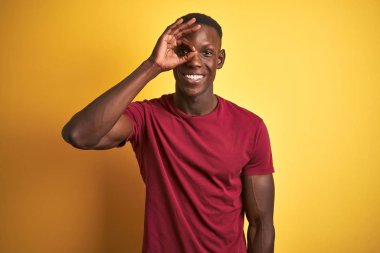 Kırmızı tişörtlü genç Afro-Amerikalı adam izole edilmiş sarı arka planda dikiliyor. El hareketleriyle gülümsüyor, gözleriyle mutlu yüzle parmaklarının arasından bakıyor..