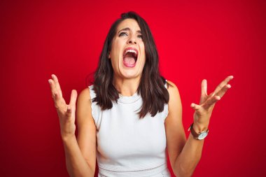 Young beautiful woman wearing white dress standing over red isolated background crazy and mad shouting and yelling with aggressive expression and arms raised. Frustration concept.