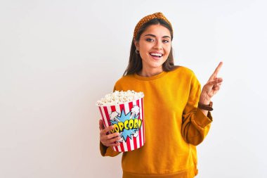 Genç ve güzel bir kadın, izole edilmiş beyaz arka planda duran patlamış mısırları tutarken mutlu bir şekilde elini ve parmağını kenarı işaret ediyor.