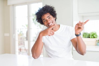 Genç Afro-Amerikalı adam, günlük beyaz tişörtler giyip evde oturmuş gülümsüyor ve kameraya bakıyor. İki eliyle ve parmaklarıyla işaret ediyor..