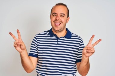 Sıradan çizgili polo giyen genç adam izole edilmiş beyaz arka planda dikiliyor ve iki elini de zafer işareti yaparken gösteren bir dille gülümsüyor. İki numara..