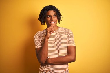 Rastalı tişörtlü Afro bir adam izole edilmiş sarı arka planda duruyor. Elleri çenesinde soru sorarken, düşünceli bir ifade takınıyor. Gülen ve düşünceli bir yüz. Şüphe kavramı.