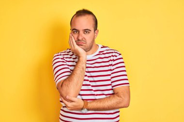Sıradan çizgili tişört giyen genç bir adam izole edilmiş sarı arka planda dikilirken kolları çaprazlanmış depresyondan sıkılmış ve yorgun görünüyor..