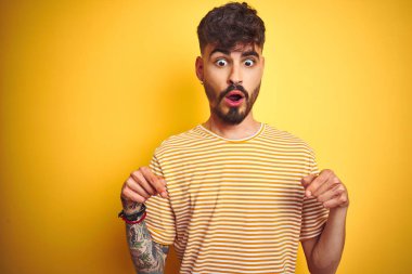 Dövmeli, çizgili tişörtlü genç adam izole edilmiş sarı arka plan üzerinde duruyor parmaklarıyla reklamı gösteriyor, şaşırmış yüz ve ağzı açık.