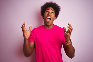 Afro saçlı genç Amerikalı adam pembe arka planda duran tişörtlü çılgın ve sinirli bir şekilde bağırıyor ve agresif bir ifadeyle kollarını kaldırıyor. Hayal kırıklığı konsepti.