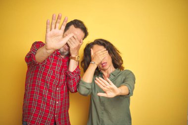 Güzel bir orta yaşlı çift, izole edilmiş sarı arka planda, elleriyle gözleri kapatıyor ve üzgün ve korku ifadeleriyle jest yapıyor. Utanç verici ve olumsuz bir kavram..