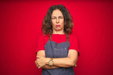Kırmızı izole edilmiş arka plan şüphecisi ve gergin, kollarını çapraz yapmış, onaylanmayan yüz ifadesi üzerine önlük giyen orta yaşlı bir kadın. Negatif kişi.