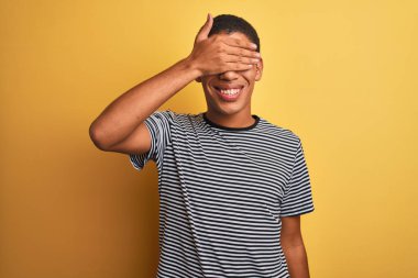 Genç yakışıklı Arap adam izole edilmiş sarı arka planda donanma tişörtü giyiyor. Gülümsüyor ve elleri yüzünde şaşkınlıktan gözleri kapatıyordu. Kör kavram.