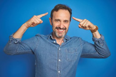 Yakışıklı, orta yaşlı, gri saçlı, mavi arka planda gülümseyen, iki eliyle başını gösteren, harika bir fikir ya da düşünce, iyi bir hafıza.