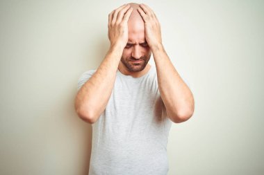 Sakallı genç ve kel bir adam izole edilmiş arka planda günlük beyaz tişört giyiyor. Ağrı ve migren yüzünden baş ağrısı ve stres altında. Ellerini başının üstüne koy..