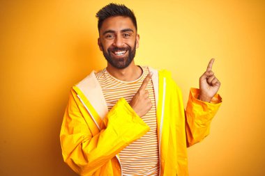 Yağmurluk giyen genç Hintli adam izole edilmiş sarı arka planda duruyor gülümsüyor ve kameraya bakıyor iki eliyle ve parmaklarıyla kenarı işaret ediyor..