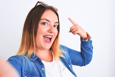 İzole edilmiş beyaz arka planda duran kot gömlek giyen genç ve güzel bir kadın mutlu bir şekilde elini ve parmağını gösterdi.