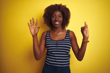 İzole edilmiş sarı arka plan üzerinde çizgili tişört giyen Afrikalı genç bir kadın yedinci parmaklarını gösterirken kendinden emin ve mutlu bir şekilde gülümsüyor..