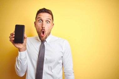 Genç ve yakışıklı iş adamı sarı arka plan üzerinde akıllı telefon ekranı gösteriyor. Şaşırmış bir yüzle korkmuş ve korku ifadesinden heyecanlanmış.